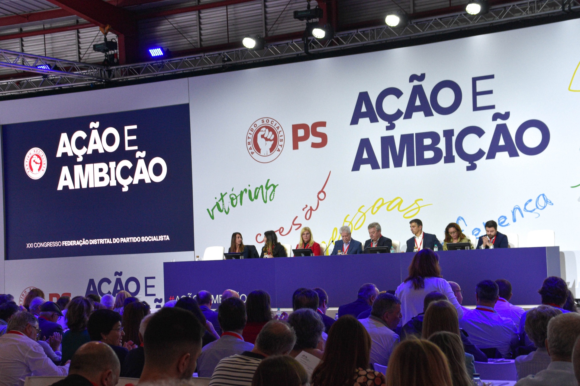 Mesa de trabalhos no palco do congresso com participantes em sessão.