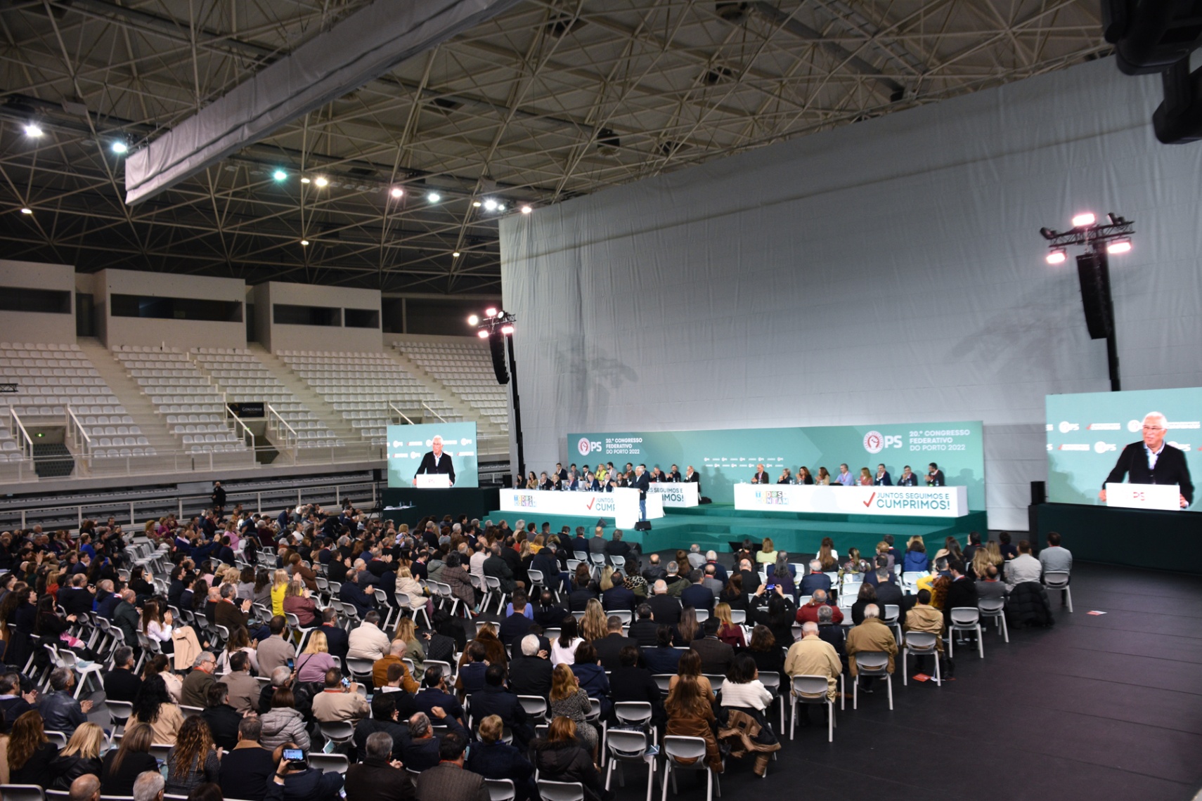 Plano geral do recinto do congresso com palco central e público distribuído pela sala.